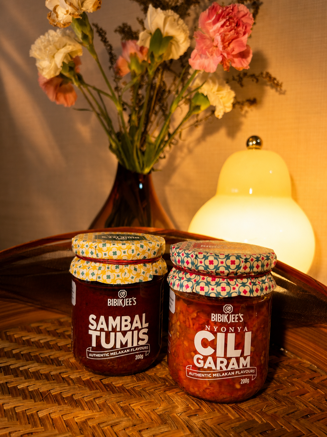 Two jars of Bibikjee's authentic cili garam and sambal on a woven tray, with a cozy home ambience